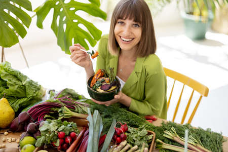 Portrait of a young cheerful woman eats vegetarian bowl while sitting by the table full of fresh vegetables, fruits and greens indoors. Healthy natural eating and lifestyle conceptの写真素材