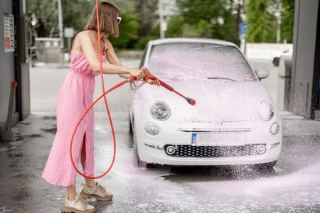 Young woman in pink dress washing her tiny car with nano foam at car wash during summer time. Easy...