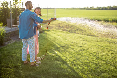 Lovely couple watering lawn at backyard of their country house on sunset. Young family taking care of their garden spending summer time togetherの写真素材