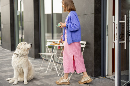 Young stylish woman goes with coffee cup from a coffee shop, cute white dog waits for her near the...