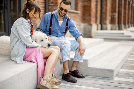 Young stylish couple sit together and have fun with their white dog on a street. Young hipsters hang out together near office outdoorsの写真素材