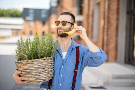 Portrait of weird stylish businessman holds green plant and banana near his ear, like talking on phone outdoors. Cool guy wearing blue shirt and suspendersの写真素材
