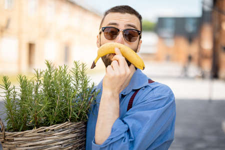 Portrait of weird stylish businessman holds green plant and banana like mustache outdoors. Cool guy wearing blue shirt and sunglassesの写真素材