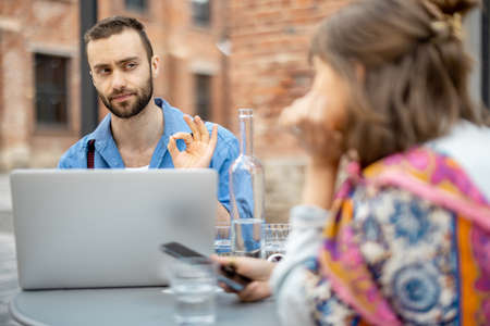 Stylish man talks with his female colleague while sitting with laptop at cafe terrace outdoors. Business talk in relaxed atmosphereの写真素材