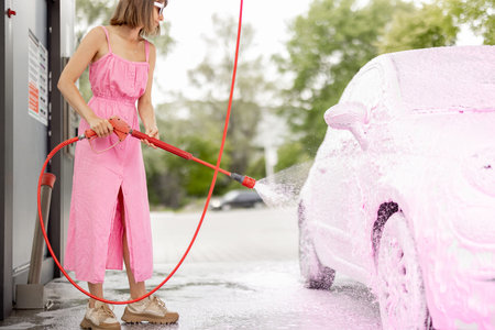Young woman in pink dress washing her tiny car with nano foam at car wash during summer time. Easy...