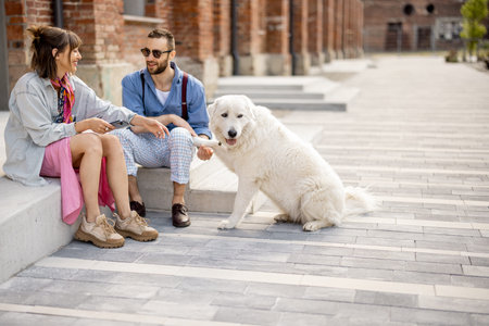 Young stylish couple sit together and have fun with their white dog on a street. Young hipsters hang...