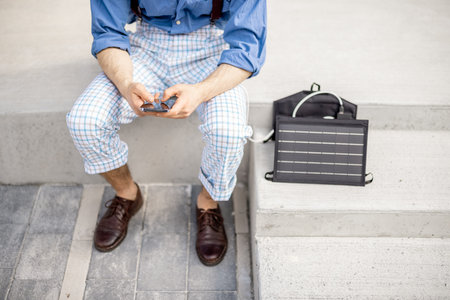 Stylish man use smart phone and charge it from portable solar panel while sitting on street...