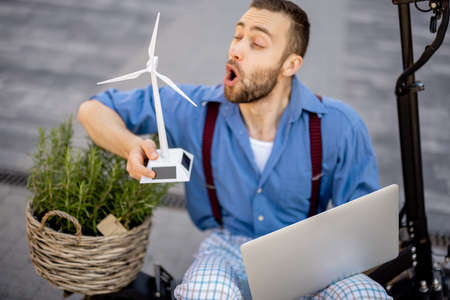 Man blows on toy wind generator while sitting with laptop and green plant on electric scooter outdoors. Concept of sustainable lifestyle and renewable energyの写真素材