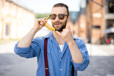 Portrait of weird stylish businessman holds banana near his ear, like talking on phone outdoors. Cool guy wearing blue shirt and suspendersの写真素材