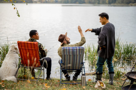 Asian man greets with his male multiracial friends fishing on river or lake coast. Concept of leisure, hobby and weekend in nature. Idea of friendship. Autumn day. View from the backsideの写真素材