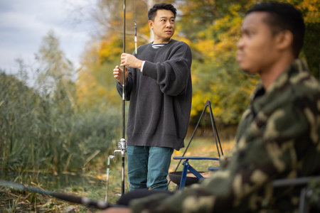 Two multiracial male friends fishing in nature. Men resting and spending time together on river or...