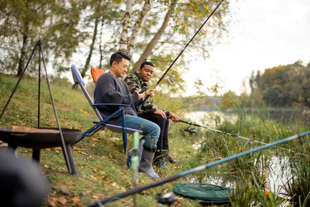 Two multiracial male friends fishing in nature. Men resting and spending time together on river or...