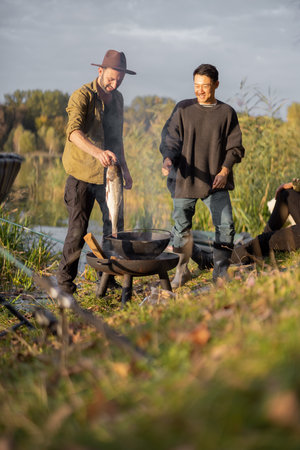 Joyful caucasian man putting big fish in cauldron for cooking Ukha while multiracial friends looking this. Men resting and fishing on lake coast at autumn. Leisure and weekend in nature. Friendshipの写真素材