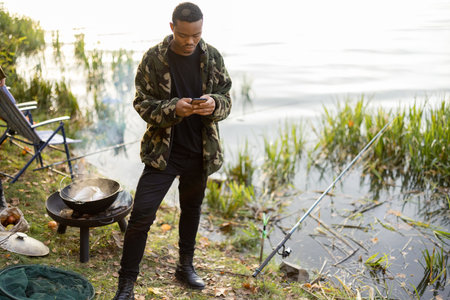 Hispanic men using smartphones during resting on river or pond coast. Men fishing and cooking on river coast at autumn. Leisure and weekend in natureの写真素材