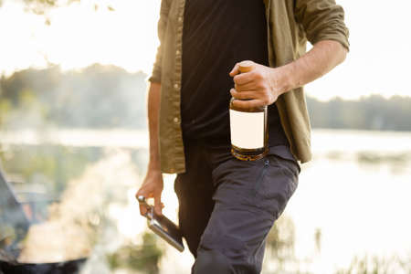Man holding bottle with alcohol while fishing on lake, close-up on bottle with blank label to copy paste. Concept of leisure in nature with alcoholic beveragesの写真素材