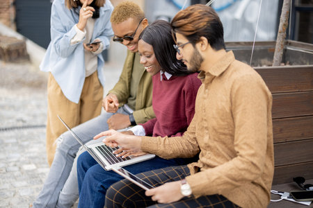 Multiracial students watching something on laptop while sitting on wooden bench outdoors. Concept of remote and e-learning. Idea of students lifestyle. Young guys and girls at university campusの写真素材