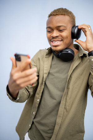 Young black man listening music in earphones and using smartphone. Handsome enjoying guy in glasses wearing jacket and t-shirt. Blue backgroundの写真素材