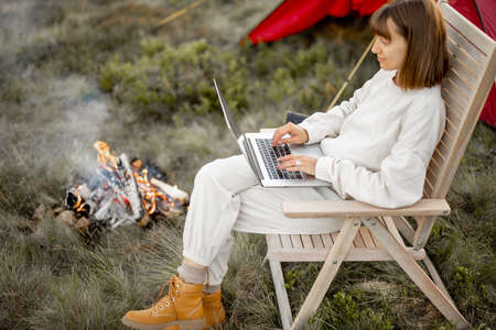 Young woman works on laptop while sitting relaxed on chair by the campfire, traveling with tent on nature. Concept of remote workの写真素材