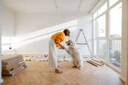 Young woman plays with her dog in room while making repairing in apartment. Fun during house renovation and friendship with pets conceptの写真素材
