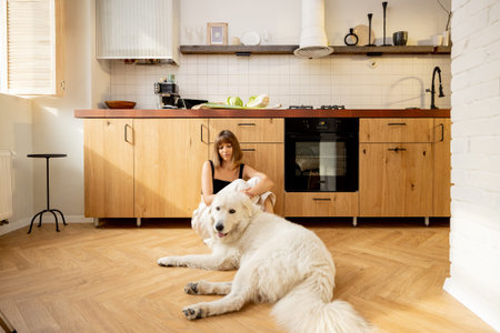 Young woman sitting relaxed with her white dog on a floor in kitchen, spending leisure time at...