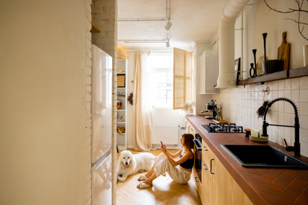 Woman uses smart phone while sitting with her dog in kitchen. Interior view on modern and stylish kitchen in beige tones. Domestic lifestyle conceptの写真素材