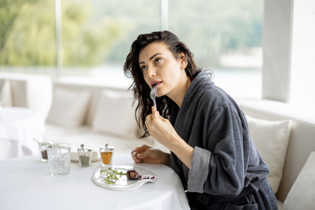 Beautiful woman in bathrobe enjoys delicious meal, sitting in white restaurant of haught cuisine during vacations at luxury spa resortの写真素材