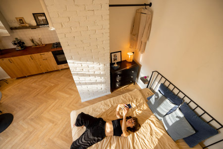 Woman lying relaxed with phone on bed in sunny studio apartment, view from above