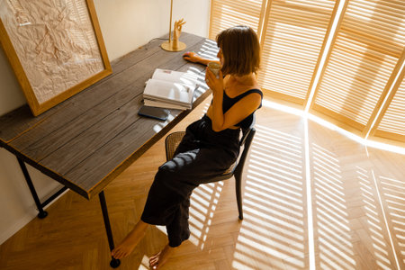 Woman reads some magazine while sitting at cozy workplace by the wooden table at home. Living room...