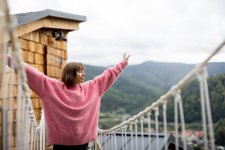 Woman in pink sweater walks on rope bridge of wooden house highly in mountains. Recreation and escaping to nature conceptの写真素材