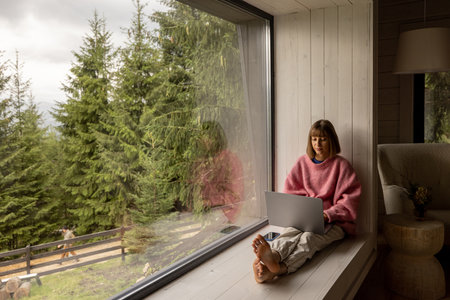 Woman works on laptop while sitting on a window with great view on mountains. Remote work from cozy place and escaping to nature conceptの写真素材