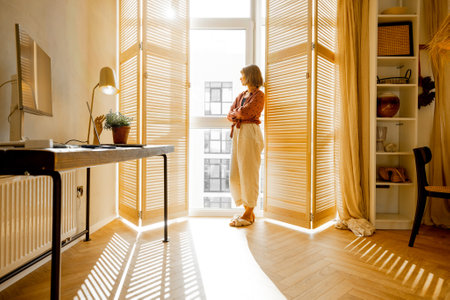 Young woman stands alone by the window blinds at cozy and sunny living room of modern apartment in beige tones. Quarantine, loneliness and life at homeの写真素材