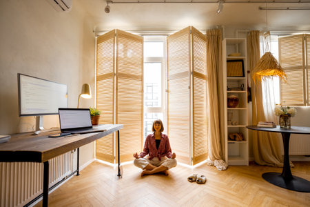 Young woman meditates after work, sitting on the floor at cozy home office. Stylish interior with workplace and computers in front. Mental health and relaxation, working from home conceptの写真素材