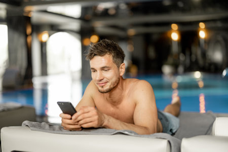 Man rests by the swimming pool indoors, talking on phone while lying relaxed on lounge bed indoors. Man spends holidays at spa resort with an indoor poolの写真素材