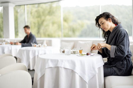 Beautiful woman in bathrobe enjoys delicious meal, sitting in white restaurant of haught cuisine during vacations at luxury spa resortの写真素材