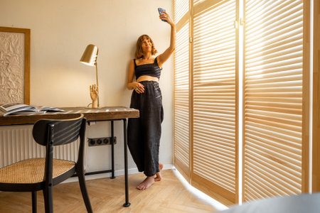 Young stylish woman in black home wear talking online or making photo on smart phone while standing near the window blinds and workplace at home. Concept of blogging and domestic lifestyleの写真素材