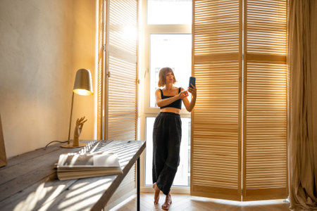 Young stylish woman in black home wear talking online or making photo on smart phone while standing near the window blinds and workplace at sunny room. Concept of domestic lifestyleの写真素材