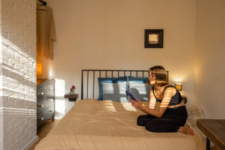 Woman using smart phone while sitting relaxed on bed in beautiful and sunny bedroomの写真素材
