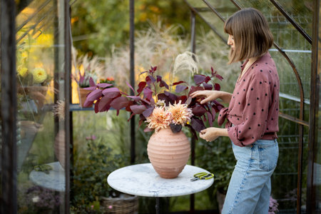 Woman florist makes a beautiful and lush bouquet in vase on a table in a small greenhouse for plants at back yard. Floristry and beauty conceptの写真素材
