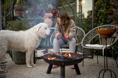 Young woman warms her hands, spending evening time with her dog by the fire at cozy and beautiful backyardの写真素材