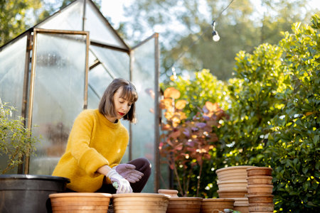 Woman planting flowers in clay jugs for growing in glass orangery at garden. Florist gardening at beautiful backyard on sunny morningの写真素材