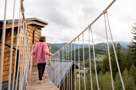 Woman enjoys scenic view on nature, while standing on the rope bridge of wooden house in mountains. Recreation and escaping to nature conceptの写真素材