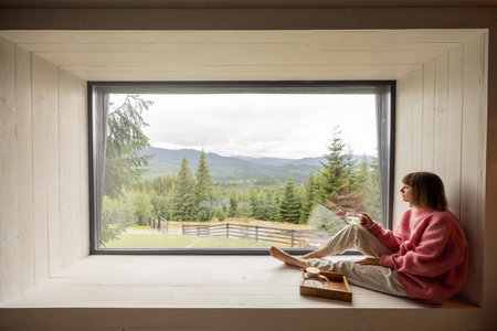 Woman sits with phone on window sill and enjoys scenic view on mountains while resting in wooden house on nature. Recreation and escaping to nature conceptの写真素材