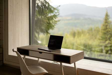 Workplace with laptop on table in front of panoramic window with scenic view on mountains and pine forest. Home office and remote work conceptの写真素材
