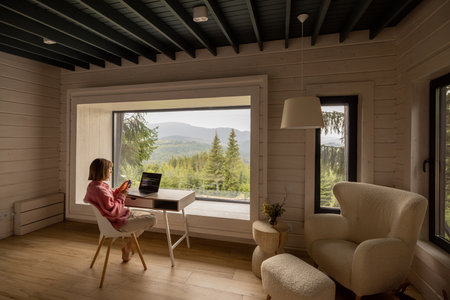 Woman works on laptop while sitting by the table in front of panoramic window with great view on mountains. Wide interior view. Remote work and escaping to nature conceptの写真素材