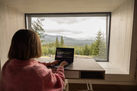 Woman works on laptop while sitting by the table in front of panoramic window with great view on...