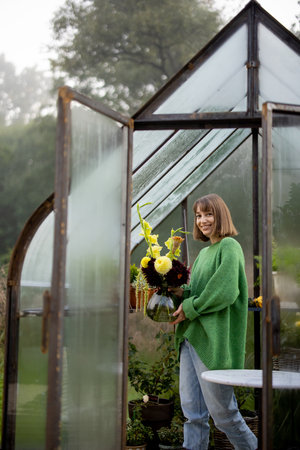 Portrait of woman florist stands with beautiful bouquet of flowers in tiny orangery at backyard. Vintage greenhouse made of rusty metal and glassの写真素材