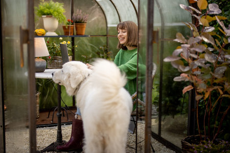 Woman plays with her dog while sitting relaxed in beautiful greenhouse for growing plants, spending leisure time in gardenの写真素材