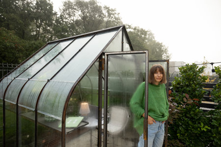 Portrait of young woman stands near greenhouse for growing plants at backyard. Gardening, leisure time in garden conceptの写真素材