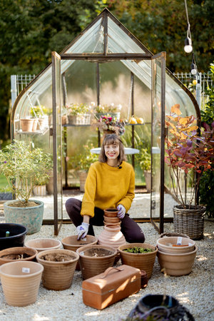 Woman plants flowers in clay jugs while sitting in front of beautiful glass orangery at garden. Florist gardening at beautiful backyard on sunny morningの写真素材