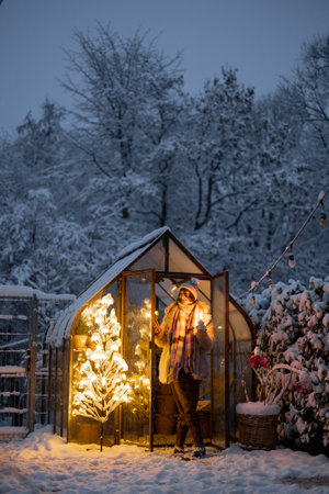 Beautiful snowy yard with vintage greenhouse and glowing tree garland. Woman with candle going out. Concept of New Year holidays and winter magicの写真素材